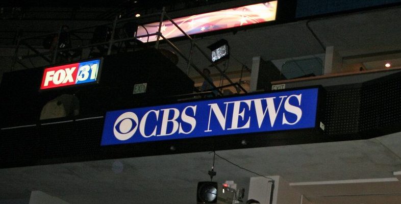 The CBS News sign displayed inside a television news studio with broadcasting equipment visible overhead.