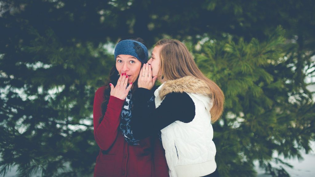 A woman whispering in another woman's ear while her hands on her lips.