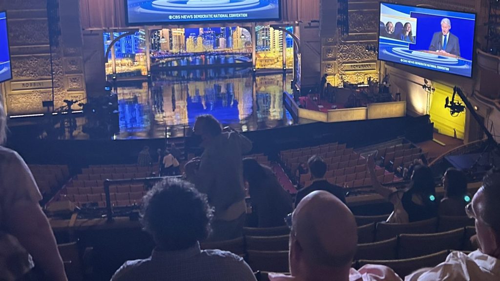 Audience members watch a large live broadcast event inside a theater featuring illuminated screens and stage production lighting.