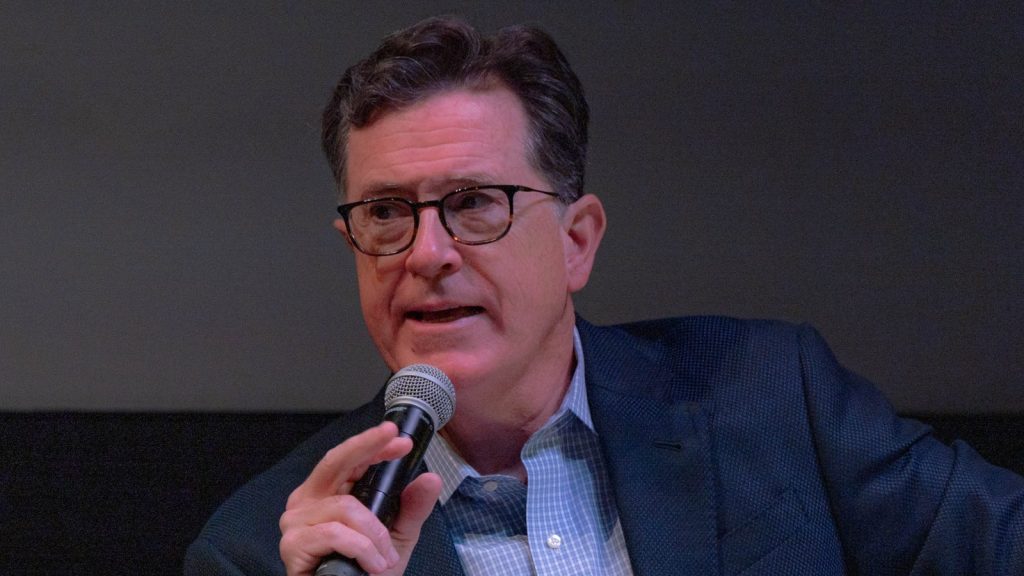 Stephen Colbert speaks into a handheld microphone during a live event.