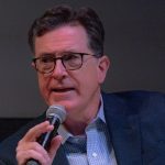 Stephen Colbert speaks into a handheld microphone during a live event.