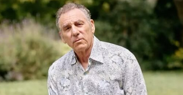 Michael Richards wearing a patterned button-up shirt while posing outdoors with blurred greenery in the background.