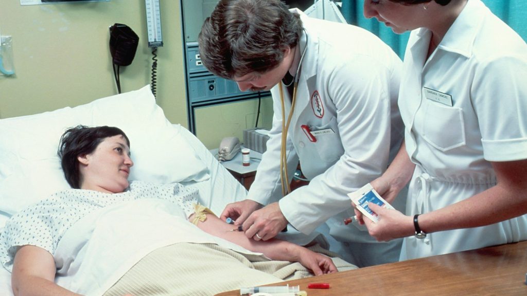 Medical staff treating a patient in a hospital bed while checking an intravenous line during care.