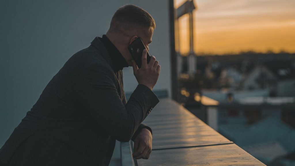 A man in a suit making a phone call on a rooftop balcony at sunset.