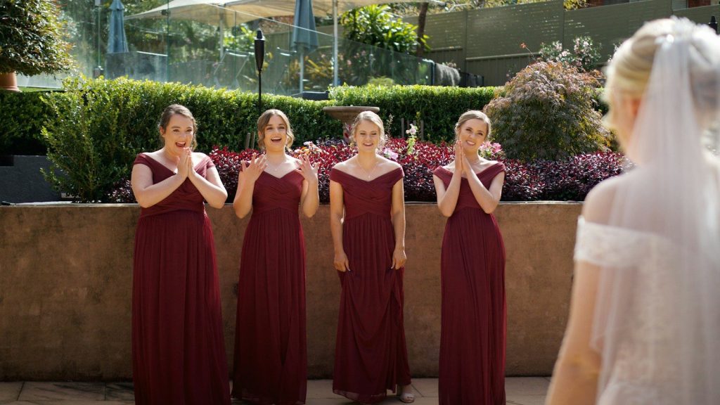 Bridesmaids in burgundy gowns reacting with excitement as bride appears at wedding ceremony.