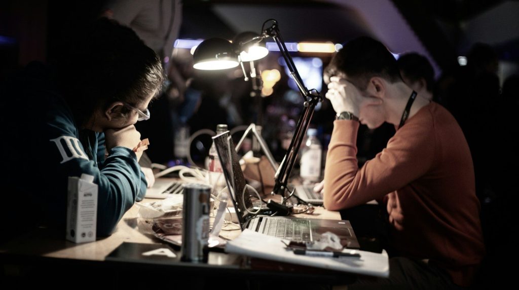 Two people working at a dimly lit desk while reviewing information on laptops under focused lighting.