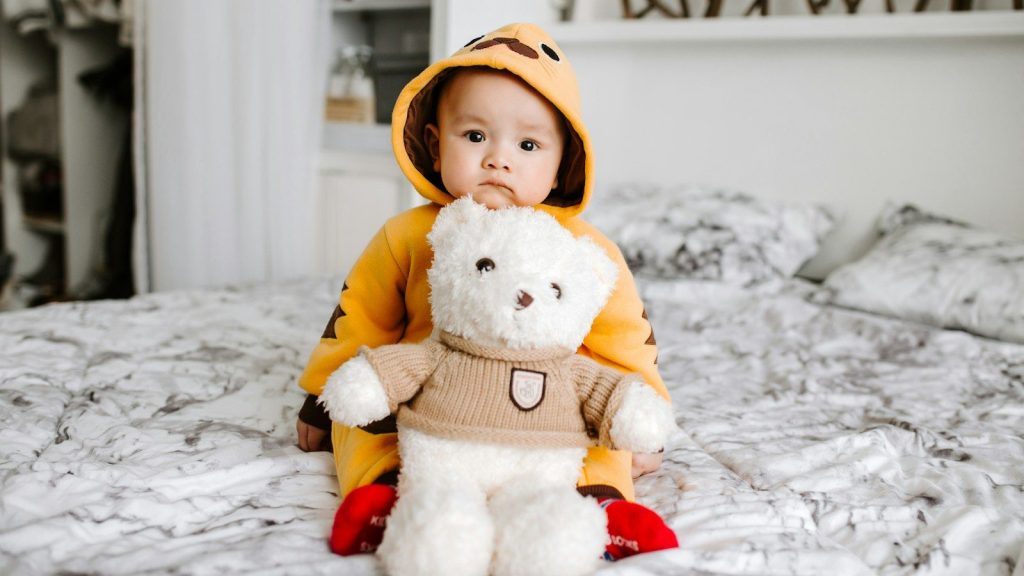 Toddler sitting on bed beside white bear plush toy.