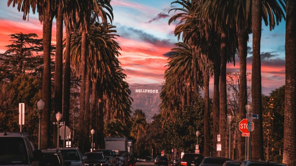 A palm-lined city street at sunset, with cars parked along the road and the Hollywood sign visible in the distance.
