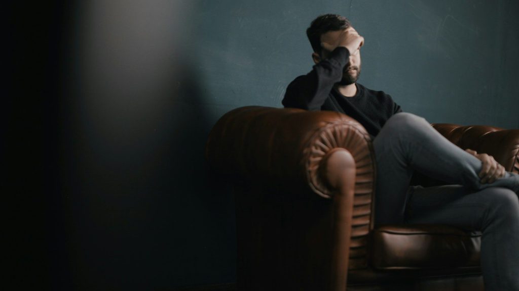 A man holds his head while sitting on a sofa.