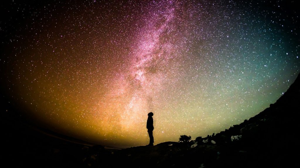 Silhouette of a man standing beneath a vibrant, star-filled night sky with the Milky Way stretching overhead.