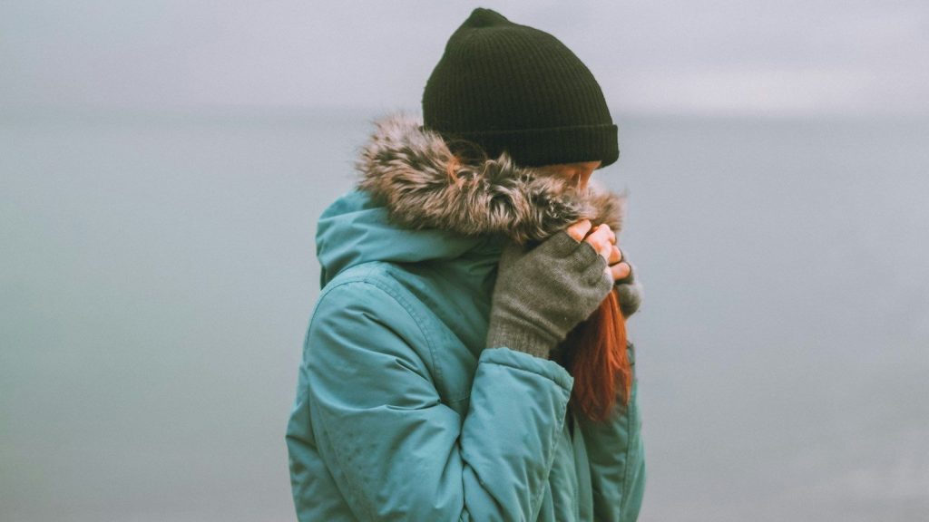 A woman in a jacket and a black knit cap covering her face with her hand.