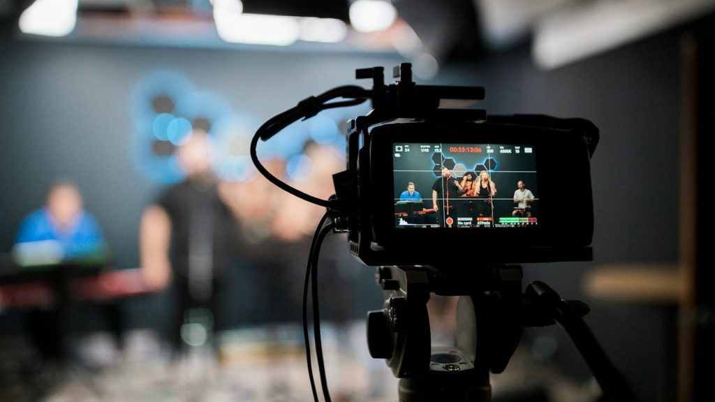 A television camera records a live studio interview with guests in the background.