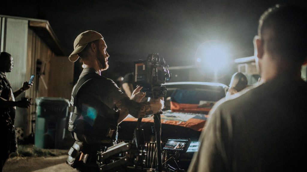 Camera operator filming a car at night on a movie set, with crew nearby.