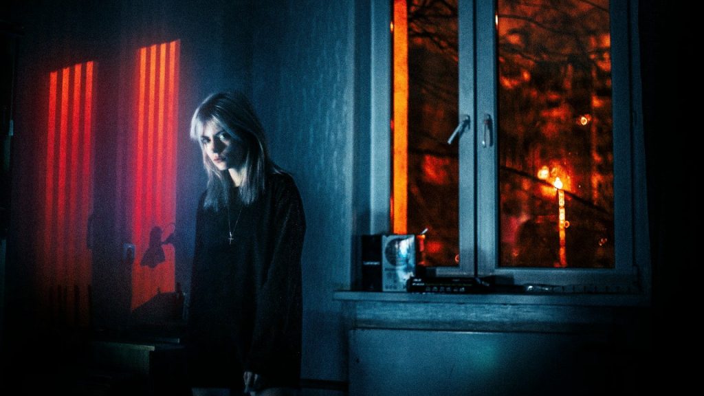 Woman standing in a dimly lit room, red light glowing through the window beside her.