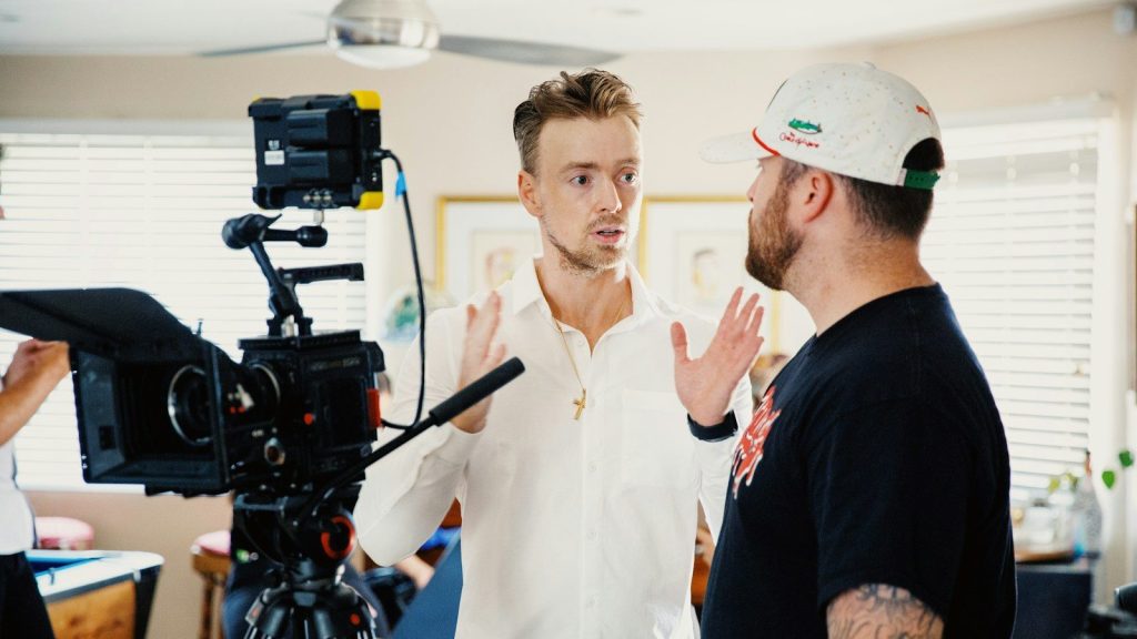 Two men discuss a film production beside a movie camera.