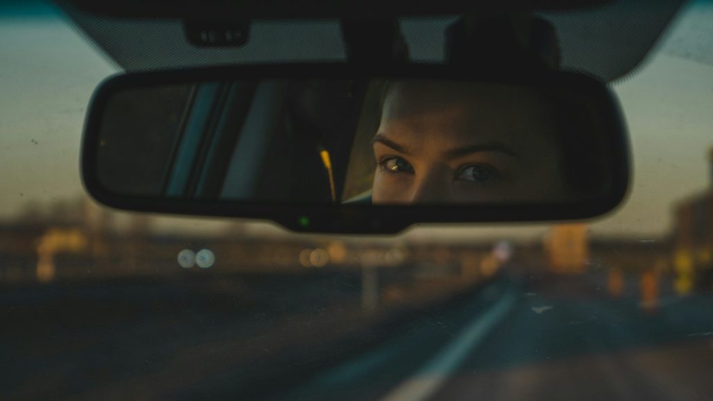Close-up of a driver's reflection in a rearview mirror on an open road.