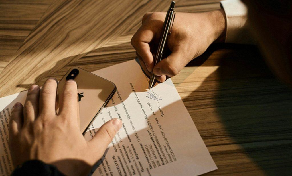Person signing a contract document with a pen on a wooden table.