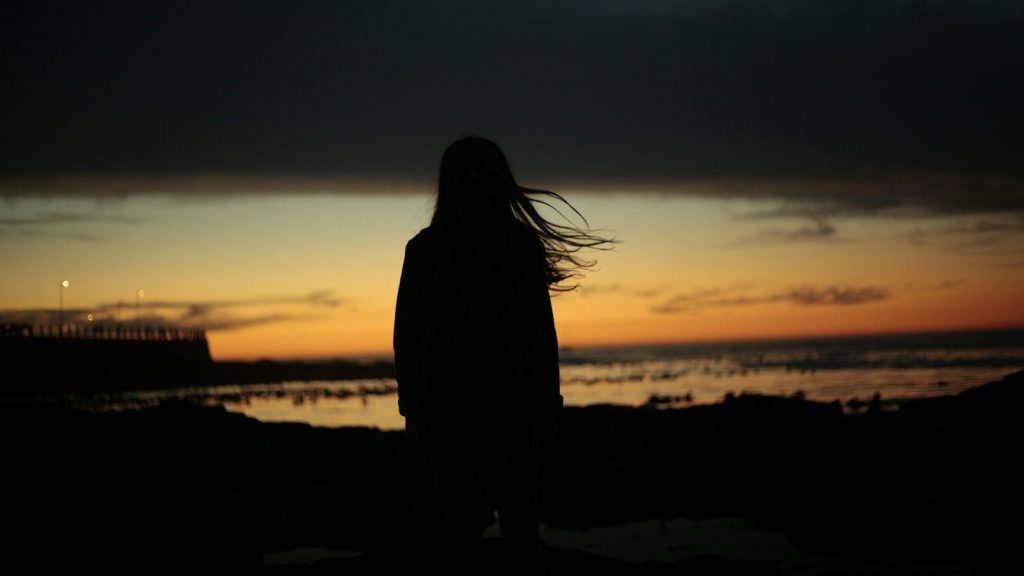 Silhouette of a woman standing by the water at sunset beneath dark clouds.