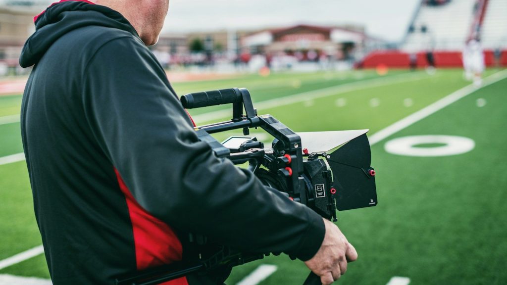 A camera operator films a football field during a live sports broadcast.